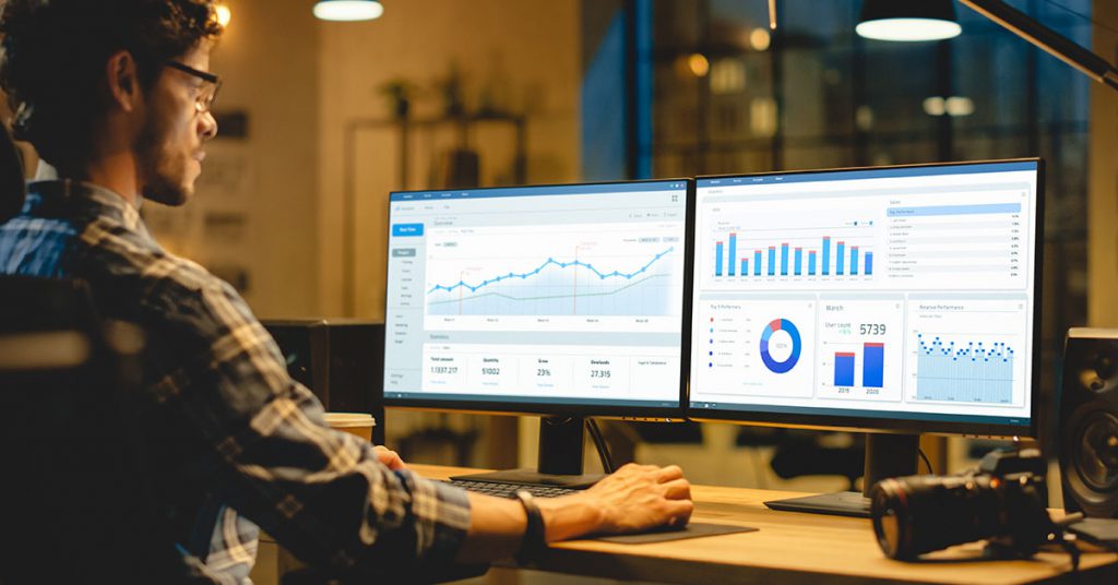 Man sitting at a desk with two monitors showing analytics dashboards