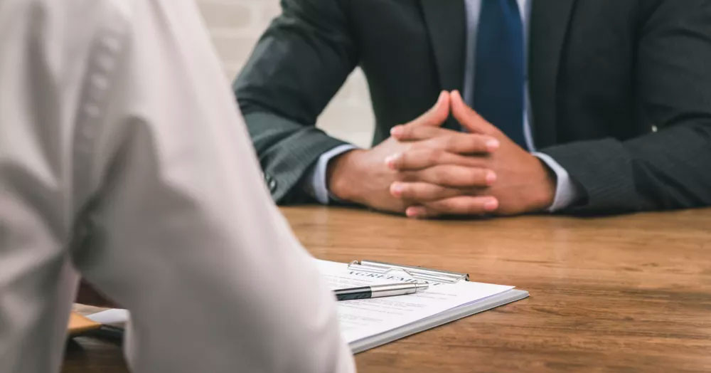 Person wearing a suit with hands folded onto a wooden desk