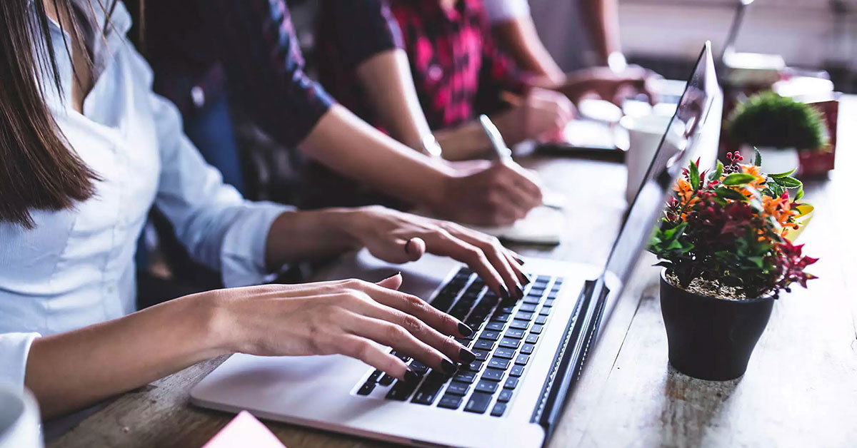 Pair of hands hovering over a keyboard while other people take handwritten notes