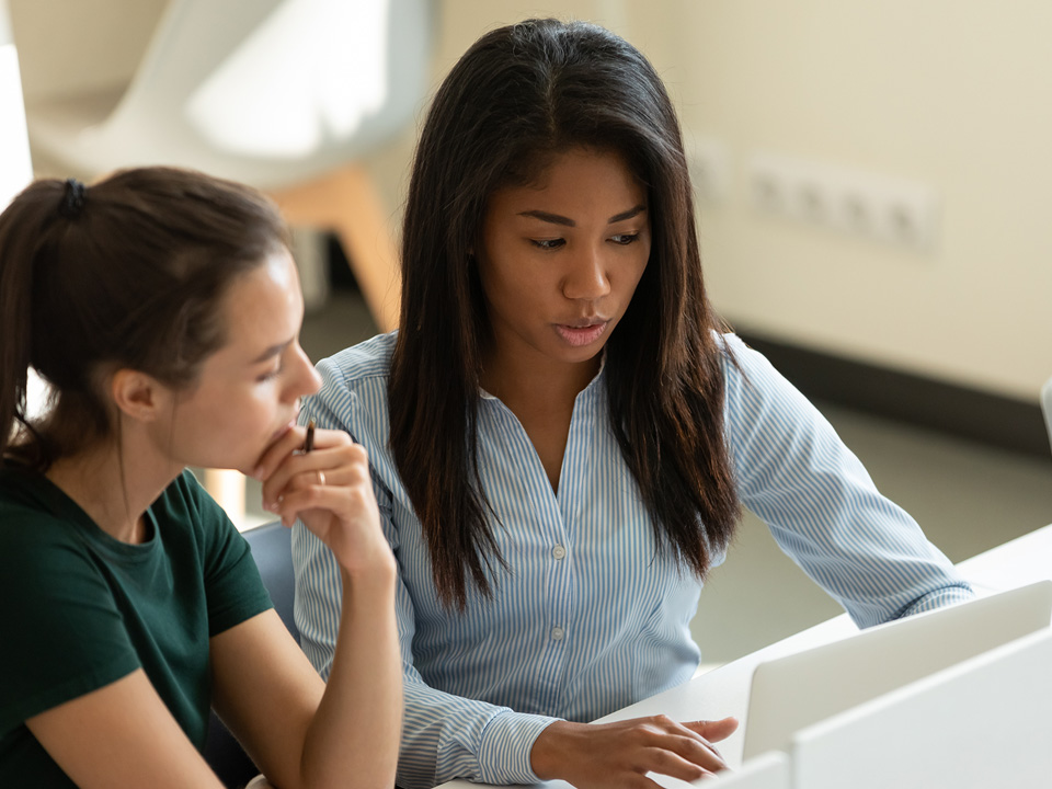 Serious diverse multiethnic student girls, interns collaborating on task at laptop together. Corporate mentor supervising new hired employee, helping with work task. Coworkers discussing project